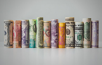 Rolled banknotes from various currencies, including the US dollar and Euro, arranged in a row against a neutral background.